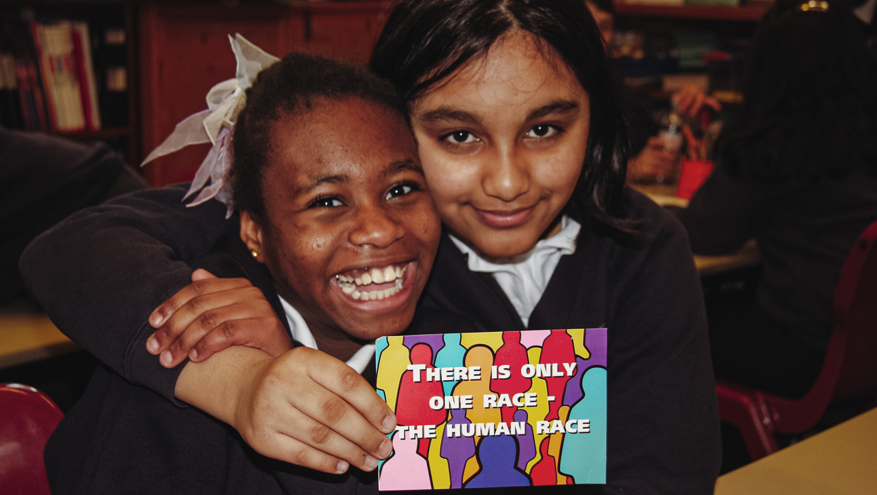 Two schoolgirls in a classroom are smiling and holding a postcard. It reads: "There is only one race - the human race."