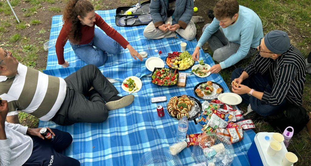 HIAS+JCORE staff, JUMP befrienders and young people sit on a blanket at the 2023 JUMP summer picnic. They are talking and eating food together.