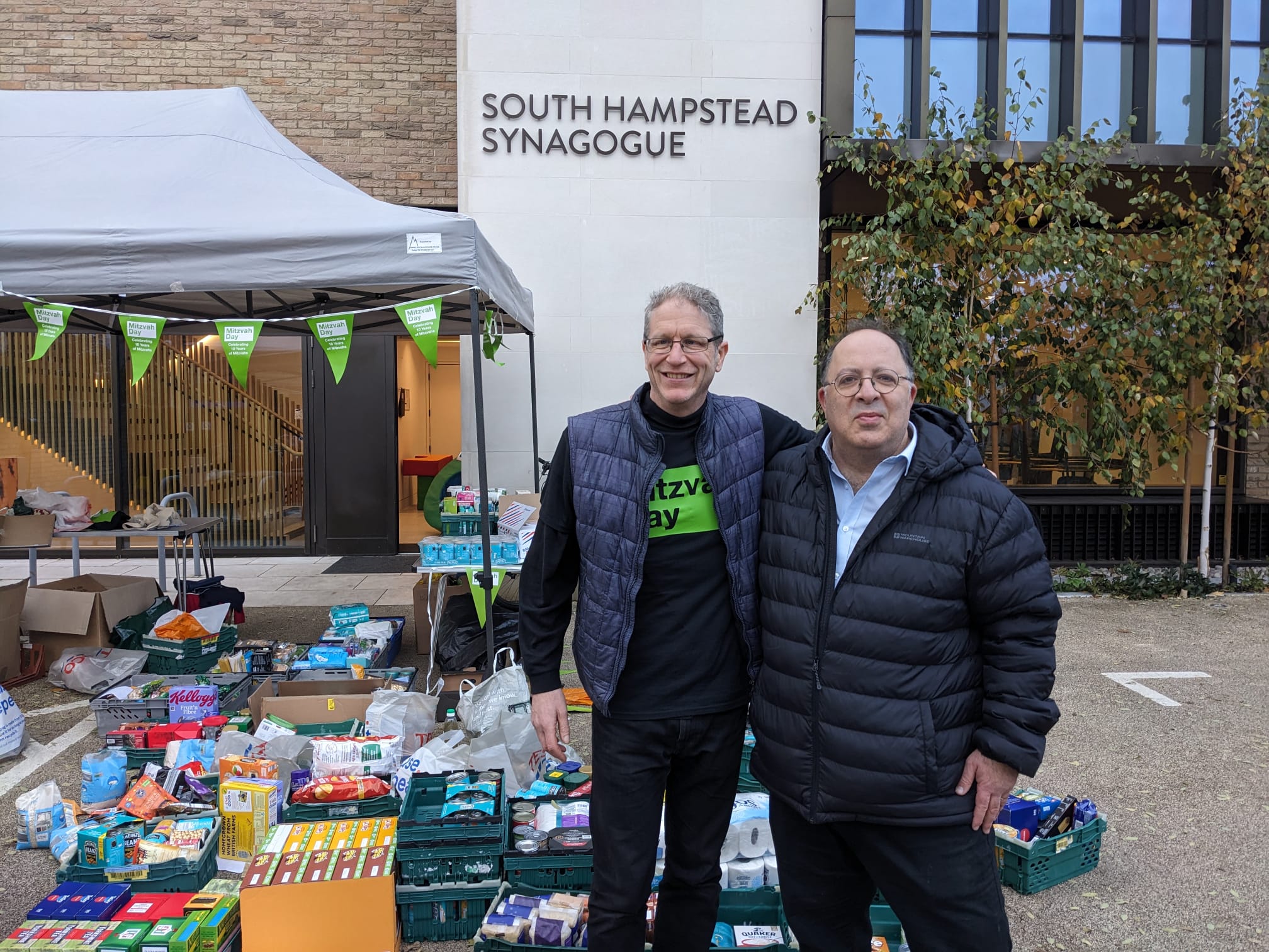 David Mason stands with a man outside South Hampstead Synagogue. They are stood in front of a collection of food and other goods, under a marquee.