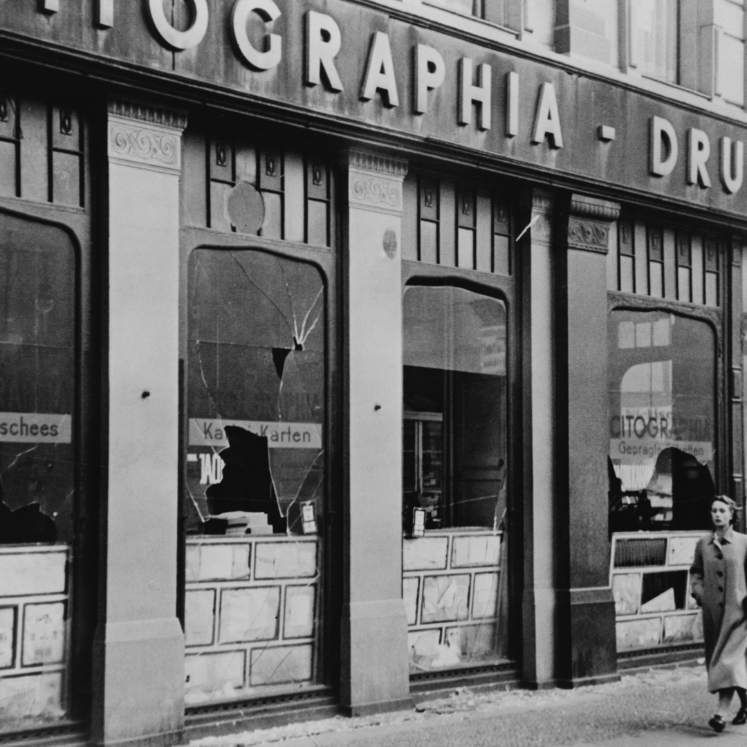 A black and white photo of a shop front damaged during Kristallnacht. Its windows have been smashed.