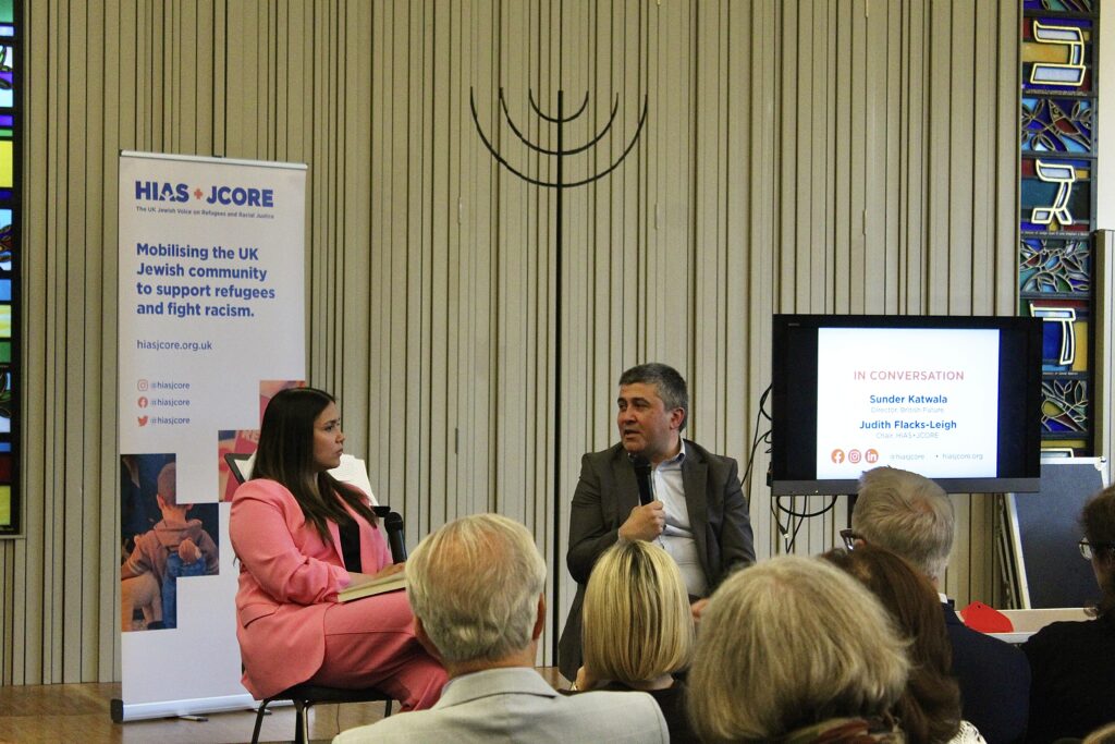 Judith Flacks (HIAS+JCORE's Chair) and Sunder Katwala (British Future Executive Director) are speaking on stage and sat down in chairs. They are in front of an audience. Sunder is sat on the right and speaking into his microphone. Judith is sat on the left and holding a microphone on her lap. Behind them is a screen displaying "In conversation. Sunder Katwala and Judith Flacks-Leigh", and a banner stand which reads: "HIAS+JCORE. Mobilising the UK Jewish community to support refugees and fight racism". The banner text is in blue on a white background.