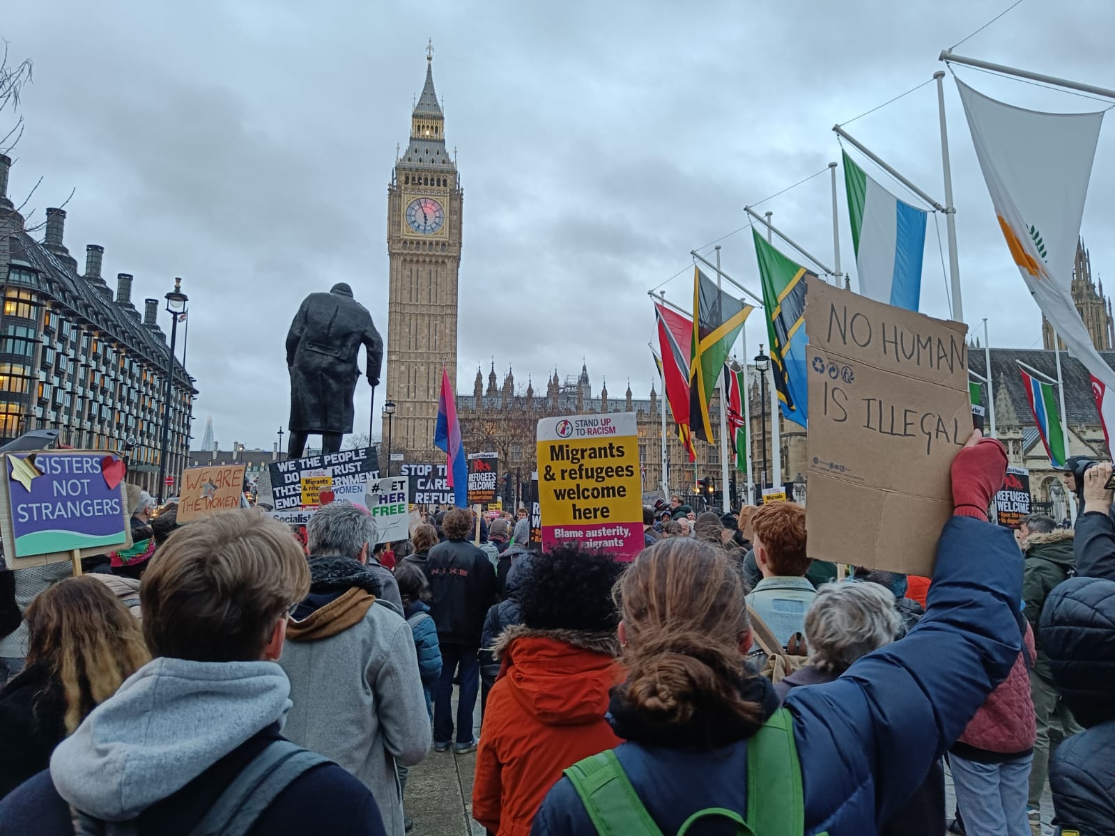 Photo shows a demonstration against the Illegal Migration Bill at Parliament Square, London. A crowd of people are stood with their backs turned to the camera. In the distance, Churchill's statue and Big Ben can be seen.