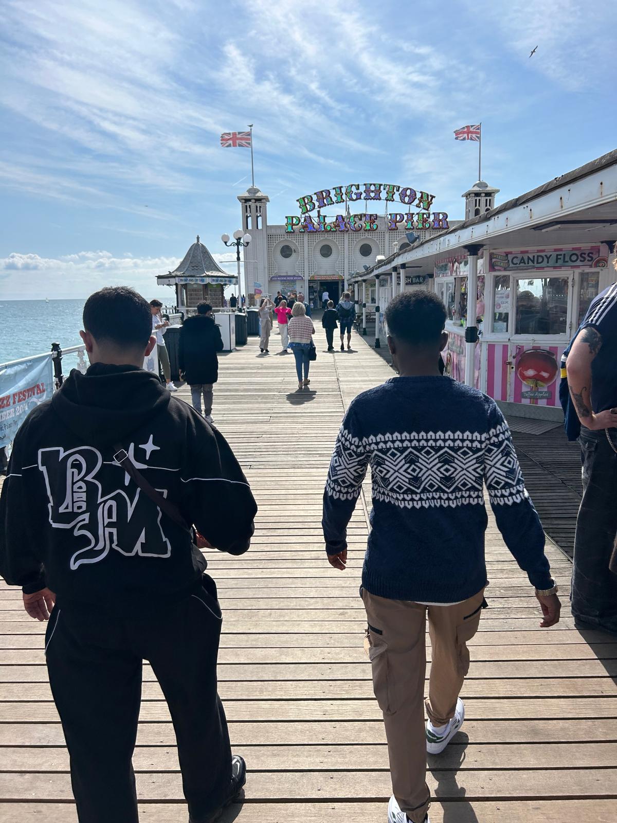 Two young males walk along Brighton pier. It is sunny.