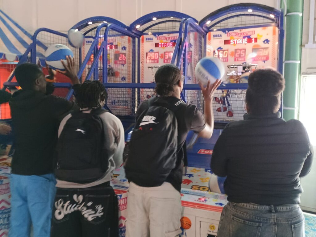 Four young people on JUMP are playing a Basketball machine. They are throwing balls into hoops.